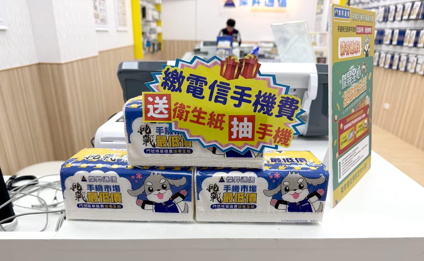 傑昇通信門市櫃檯特寫，展示「繳電信手機費送衛生紙抽手機」活動立牌與專屬包裝衛生紙贈品。